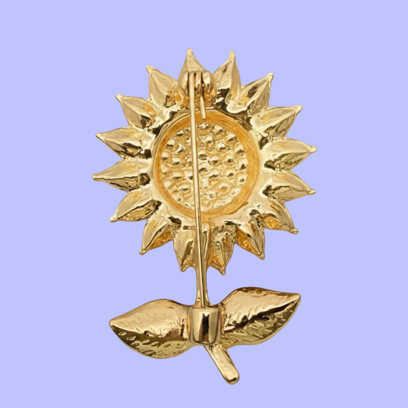Backside of sunflower-shaped brooch with yellow petals, green leaves, and a gold center against a blurred natural background