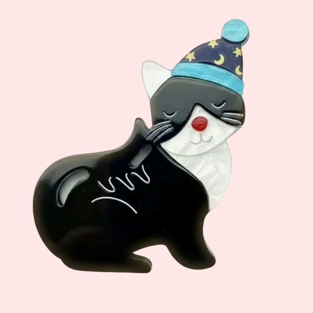 Black and white cat brooch wearing a blue nightcap with star patterns on a light peach background