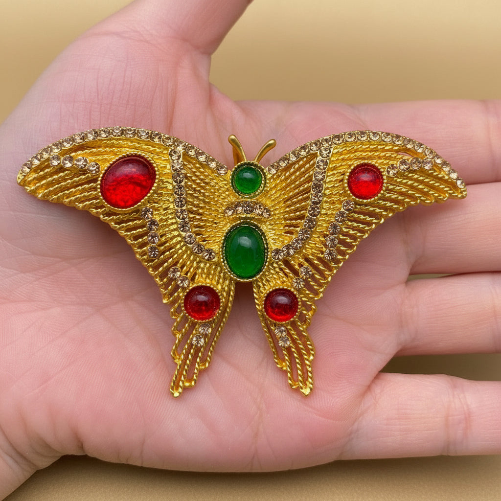 Golden butterfly brooch with red and green gemstones held in a hand against a cream colored background