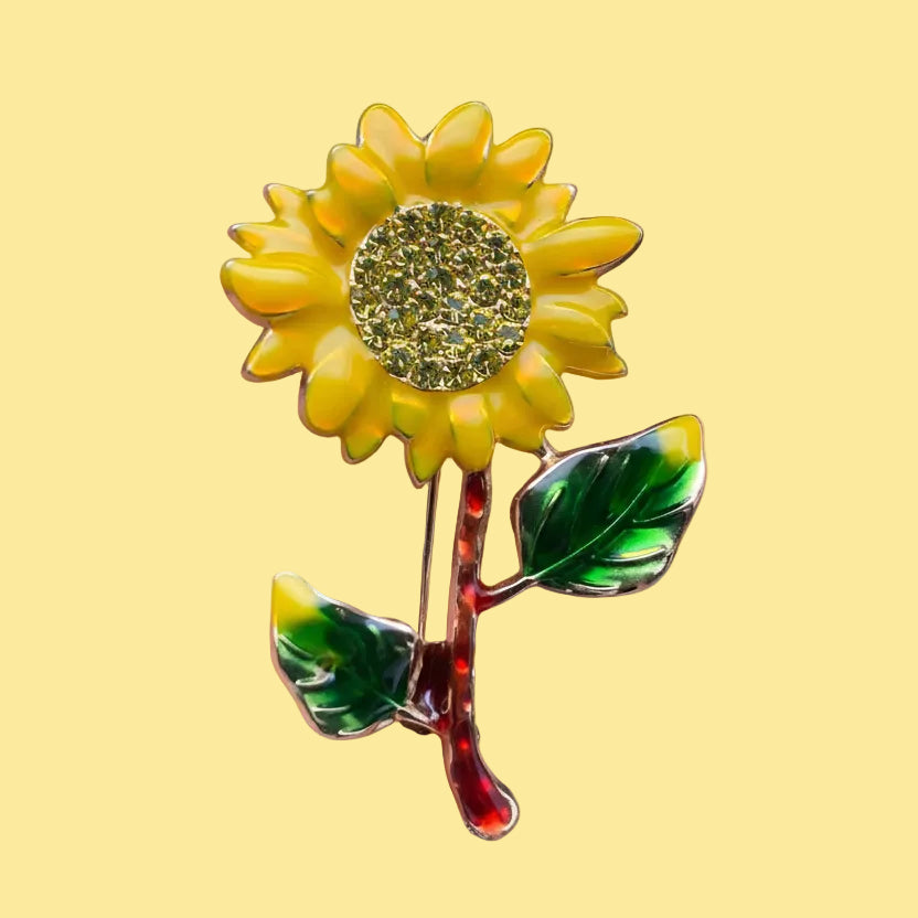 Sunflower-shaped brooch with a yellow center and green leaves on a pale yellow background