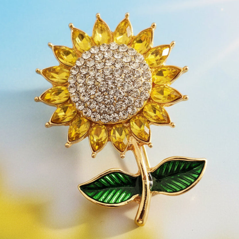 Sunflower-shaped brooch with yellow petals, green leaves, and a gold center against a blurred natural background